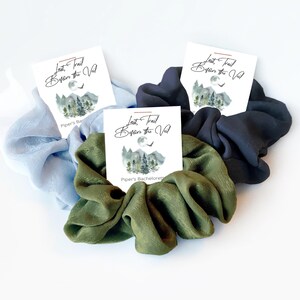 May include: Three fabric hair scrunchies in light blue, olive green, and dark charcoal gray. Each scrunchie is attached to a white card featuring a mountain and forest illustration, a moon, and the text Last Trail Before the Veil.