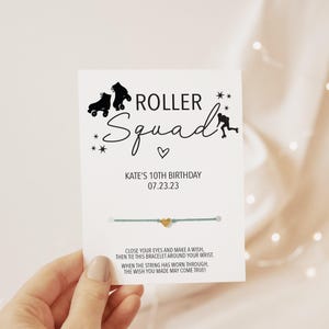 May include: A white card featuring the text ROLLER SQUAD, Kate's 10th Birthday, and 07.23.23, with black roller skate icons and stars. A teal string bracelet with a small gold heart charm is attached to the center of the card.
