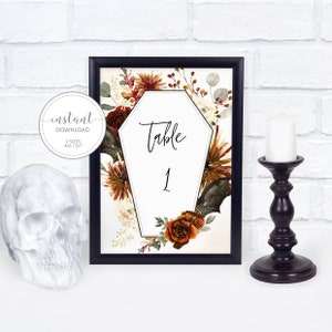 May include: A printable Halloween table number card with a black frame. The card features a white coffin-shaped design with the text "Table 1" in black. The coffin is surrounded by a floral arrangement of orange, red, and white flowers, leaves, and bats.