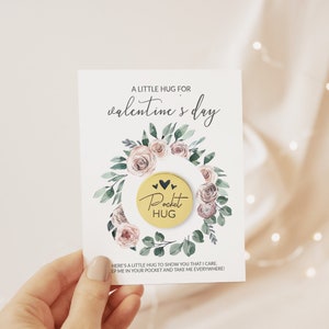 May include: A Valentine's Day card with a floral wreath design. The card features the text "A Little Hug for Valentine's Day" and a gold-colored coin that says "Pocket Hug". The card also includes a message at the bottom.