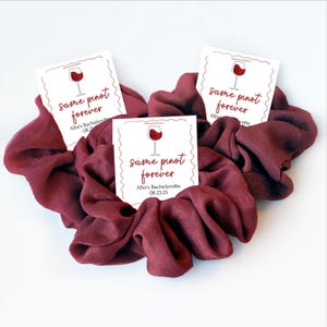 May include: Burgundy scrunchies with three white cards. Each card has a wine glass illustration and the text "same pinot forever" with "Allie's Bachelorette 08.23.25". The scrunchies are arranged in a heart shape.