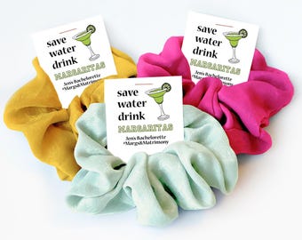 Peut inclure: Plusieurs chouchous en tissu de couleurs jaune, rose et bleu clair. Chaque chouchou est accompagné d'une petite carte blanche avec le texte "save water drink MARGARITAS" et une illustration de margarita. Les cartes indiquent également "Jen's Bachelorette #Margs&Matrimony".