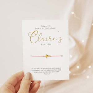 May include: A white card with gold text that reads "Thanks for Celebrating, Claire's Baptism." A pink string bracelet with a gold cross is attached. The card also includes a Bible verse. The card is held by a hand.