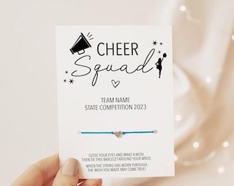 Peut inclure: Un bracelet en cordon bleu sarcelle avec une petite breloque cœur métallique fixé sur une carte de présentation blanche. La carte porte le texte noir CHEER SQUAD, TEAM NAME, STATE COMPETITION 2023 et un poème, avec des icônes de mégaphone.