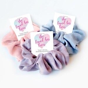 May include: Three fabric hair scrunchies in light pink, lavender, and light blue. Each scrunchie is attached to a white card featuring a watercolor heart graphic and the text "she found her lover" with "Taylor's Version #InHerBacheloretteEra".