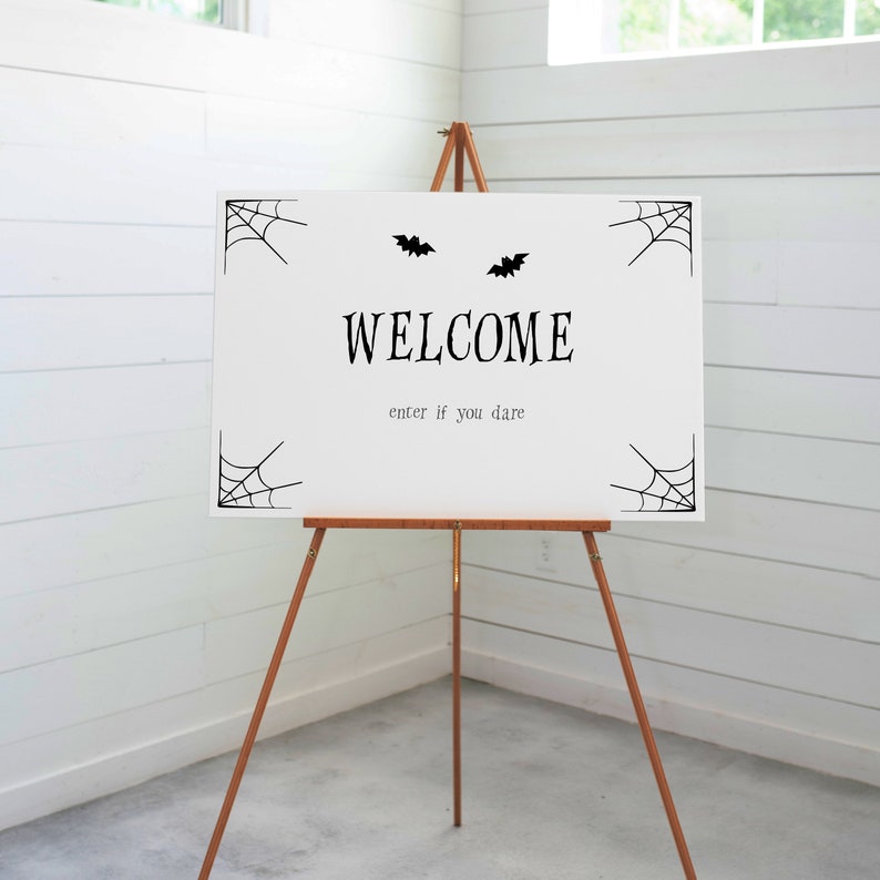 May include: A white sign on a wooden easel with the word "WELCOME" in black, accompanied by bat and spiderweb graphics. The phrase "enter if you dare" is below the main text, creating a spooky Halloween theme.