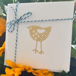 May include: A stack of white cards tied with teal and white twine. Each card features a gold bird illustration. The cards are surrounded by a sunflower and greenery.