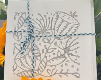May include: A stack of white greeting cards with a floral design in gray ink, tied with teal and white twine. The cards feature a botanical illustration of flowers and leaves. The cards are surrounded by yellow sunflowers and green foliage.