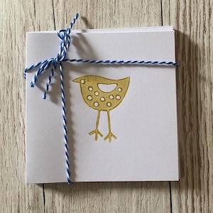 May include: A stack of white square cards tied with blue and white twine. The top card features a gold-colored bird illustration with a dotted pattern. The twine is knotted at the top, securing the cards together.