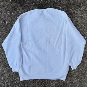 May include: A plain white sweatshirt with a crew neck and long sleeves. The sweatshirt is made of a soft material and is laid out on a textured, gray surface. The cuffs and hem are ribbed.
