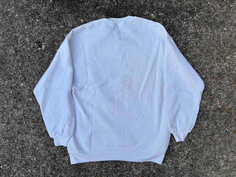 May include: A plain white sweatshirt with a crew neck and long sleeves. The sweatshirt is made of a soft material and is laid out on a textured, gray surface. The cuffs and hem are ribbed.