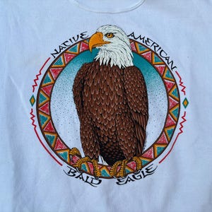 May include: White sweatshirt featuring a detailed illustration of a bald eagle. The eagle is brown, white, and yellow, set against a blue background within a circular design. The words "NATIVE AMERICAN BALD EAGLE" are printed around the circle.