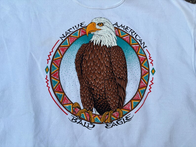 May include: White sweatshirt featuring a detailed illustration of a bald eagle. The eagle is brown, white, and yellow, set against a blue background within a circular design. The words "NATIVE AMERICAN BALD EAGLE" are printed around the circle.