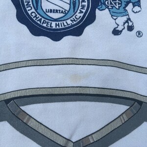 May include: White t-shirt with a blue and white University of North Carolina logo and a cartoon mascot. The logo says "University of North Carolina Chapel Hill, NC" and "Libertas".