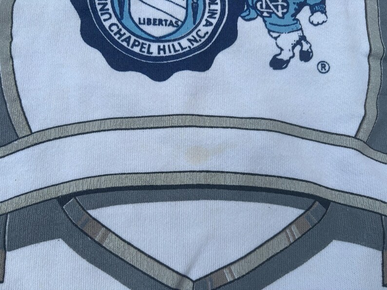 May include: White t-shirt with a blue and white University of North Carolina logo and a cartoon mascot. The logo says "University of North Carolina Chapel Hill, NC" and "Libertas".