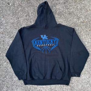 Puede incluir: Sudadera con capucha azul marino con el texto "KENTUCKY BASKETBALL" en azul y un gráfico de baloncesto. La sudadera tiene un bolsillo delantero y una capucha. La prenda está hecha de un material suave.