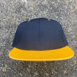 Puede incluir: Una gorra de béisbol negra y dorada con visera plana. La corona de la gorra es negra y la visera es dorada.