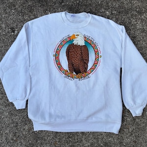 May include: White crew neck sweatshirt with a Native American bald eagle graphic. The eagle is brown with a white head and yellow beak, surrounded by a circular design with text. The sweatshirt has long sleeves and a relaxed fit.