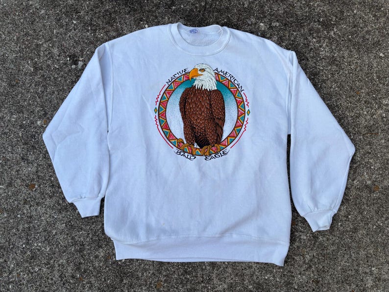 May include: White crew neck sweatshirt with a Native American bald eagle graphic. The eagle is brown with a white head and yellow beak, surrounded by a circular design with text. The sweatshirt has long sleeves and a relaxed fit.