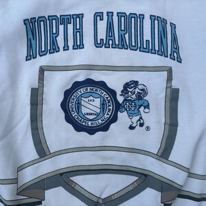 May include: White sweatshirt with a blue and gray North Carolina logo. The logo includes the words "University of North Carolina" and "Chapel Hill, NC".