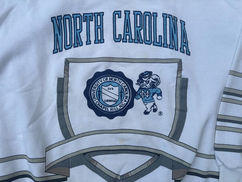 May include: White sweatshirt with a blue and gray North Carolina logo. The logo includes the words "University of North Carolina" and "Chapel Hill, NC".