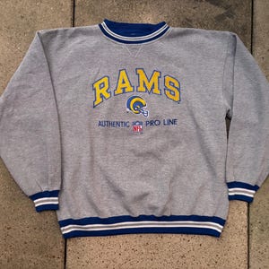 May include: A heather gray long-sleeve sweatshirt featuring the word RAMS in yellow with blue outlines, a Rams helmet graphic, and the text AUTHENTIC PRO LINE with an NFL logo. The crew neck, cuffs, and hem have blue and white striped trim.