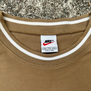 May include: A brown t-shirt with a white ribbed collar and a Nike logo tag. The tag says "XXL TTG XXG" and "Made in Peru".