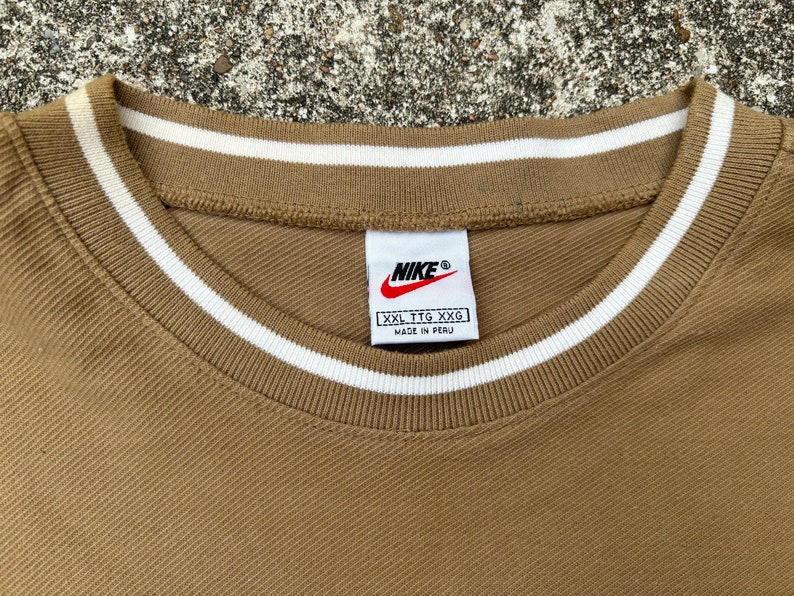 May include: A brown t-shirt with a white ribbed collar and a Nike logo tag. The tag says "XXL TTG XXG" and "Made in Peru".