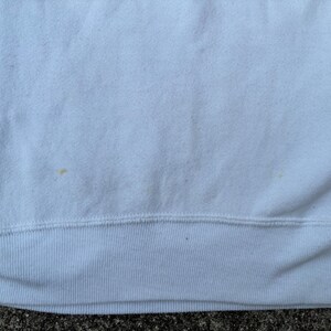 May include: Close-up of a white sweatshirt. The fabric has a textured appearance with a visible seam at the bottom. There are a few small, light-brown stains on the fabric. The bottom hem is ribbed.