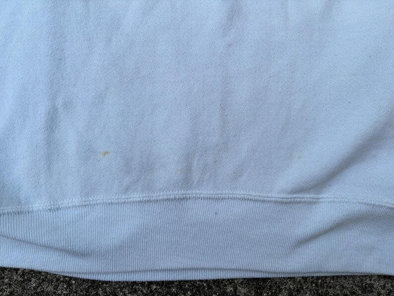 May include: Close-up of a white sweatshirt. The fabric has a textured appearance with a visible seam at the bottom. There are a few small, light-brown stains on the fabric. The bottom hem is ribbed.