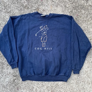 May include: Navy blue sweatshirt with a golf-themed graphic. The design features a golfer in a swing pose, with the text "COG HILL" below. The sweatshirt has a crew neck and ribbed cuffs and hem.