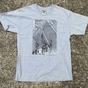 Op de afbeelding: Een grijs T-shirt met een grafische print van een groep mensen die een Amerikaanse vlag hijsen op een gebouw. De tekst "Remember 9-11-01" is onder de afbeelding gedrukt.