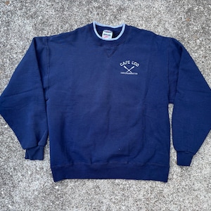 Puede incluir: Sudadera azul marino con cuello y puños azul claro. El texto "CAPE COD" está bordado en blanco sobre remos cruzados, con "MASSACHUSETTS" debajo. Prenda casual y cómoda.