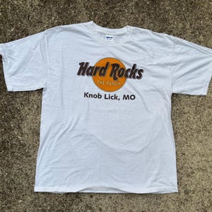Può includere: T-shirt bianca con il logo Hard Rocks, con un cerchio arancione e testo nero. Il testo recita "Hard Rocks" sopra "The Ranch" e "Knob Lick, MO" sotto. La maglietta è realizzata in un materiale morbido.