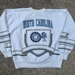 May include: White sweatshirt with a blue and gray graphic of the University of North Carolina Tar Heels logo and the text "NORTH CAROLINA".