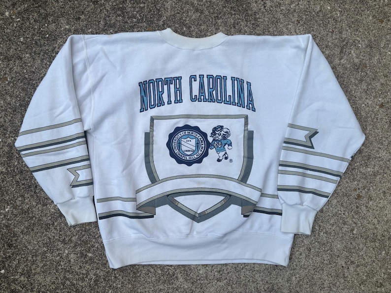 May include: White sweatshirt with a blue and gray graphic of the University of North Carolina Tar Heels logo and the text "NORTH CAROLINA".