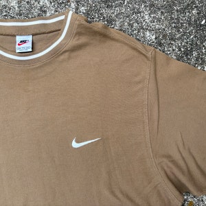 May include: A brown t-shirt with a white Nike swoosh logo on the front. The shirt has a white ribbed collar with a white stripe.