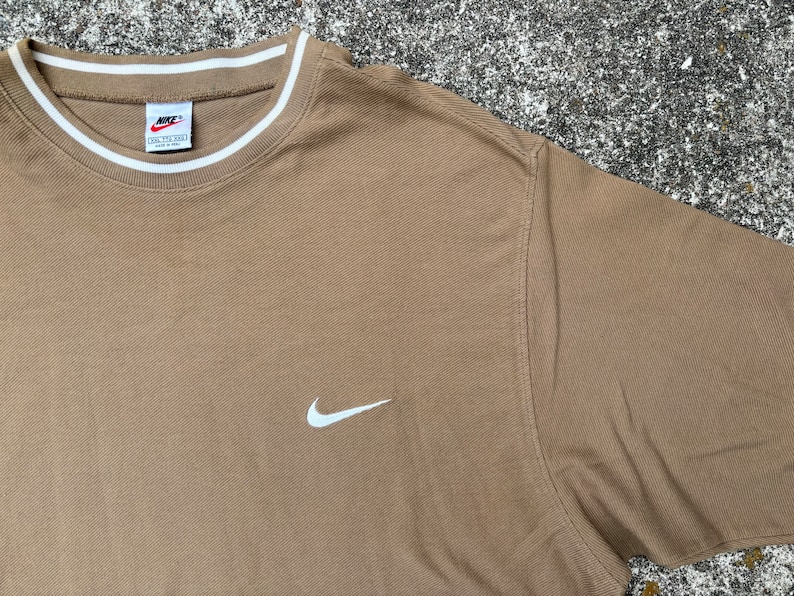 May include: A brown t-shirt with a white Nike swoosh logo on the front. The shirt has a white ribbed collar with a white stripe.
