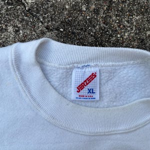 May include: Close-up of a white XL Jerzees sweatshirt. The tag is visible, with the brand name in red and the size in blue. The sweatshirt is made in the USA.