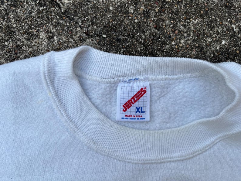 May include: Close-up of a white XL Jerzees sweatshirt. The tag is visible, with the brand name in red and the size in blue. The sweatshirt is made in the USA.