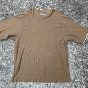 May include: A brown short-sleeved t-shirt with a white trim around the neckline and sleeves. The shirt has the word "NIKE" printed on the back in white.