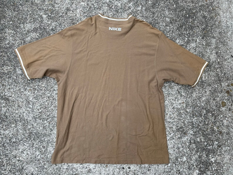 May include: A brown short-sleeved t-shirt with a white trim around the neckline and sleeves. The shirt has the word "NIKE" printed on the back in white.