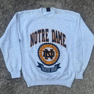 Puede incluir: Una sudadera gris claro con las palabras "NOTRE DAME" en letras arqueadas de color azul marino sobre un emblema circular. El emblema incluye las letras "ND" y las palabras "FIGHTIN' IRISH" en una pancarta azul marino.