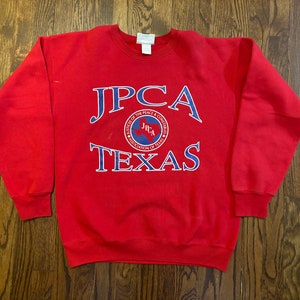 May include: Red sweatshirt with a blue and white logo that says "JPCA" and "Texas". The logo is a circle with a star and the letters "JPCA" inside.