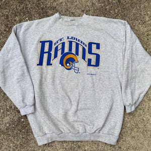 May include: A gray St. Louis Rams sweatshirt. The sweatshirt features the team name in blue block letters with a yellow outline, and a helmet graphic. The sweatshirt is made of a soft material and has long sleeves.