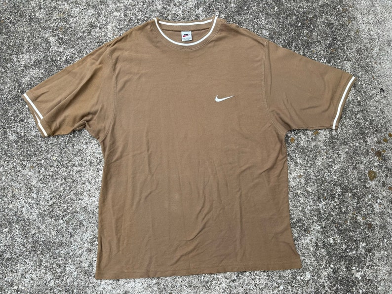 May include: A brown t-shirt with a white Nike swoosh logo. The shirt has white trim around the neckline and sleeves.