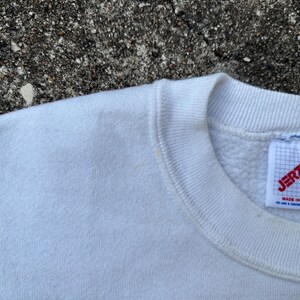 May include: Close-up of a white Jerzees XL sweatshirt. The tag is visible, with the brand name and size clearly displayed. The sweatshirt is made in the USA.