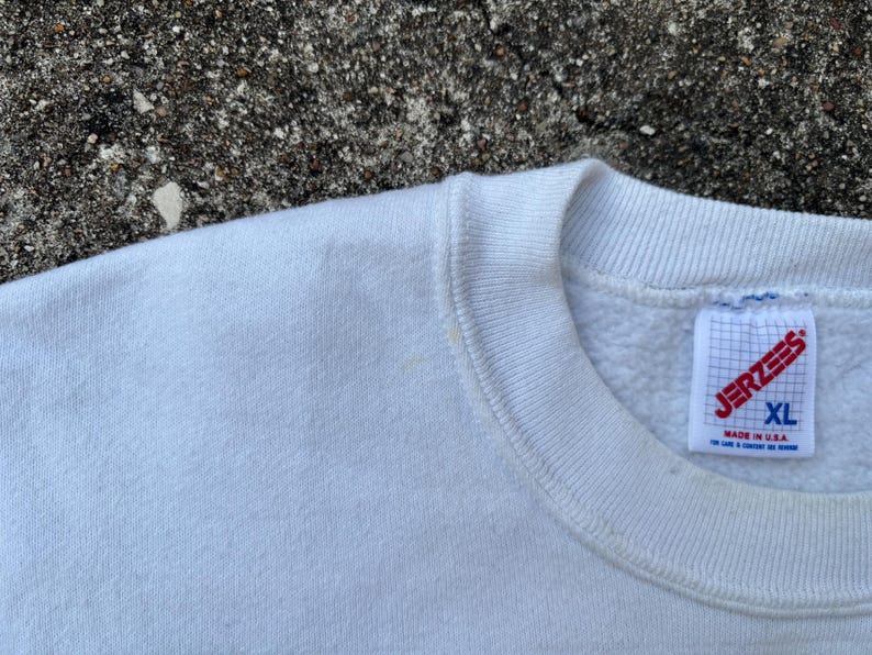 May include: Close-up of a white Jerzees XL sweatshirt. The tag is visible, with the brand name and size clearly displayed. The sweatshirt is made in the USA.