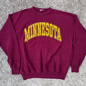 May include: A maroon sweatshirt with the word "MINNESOTA" in yellow block letters across the chest. The sweatshirt has a crew neck and long sleeves. The garment is lying flat on a textured gray surface.
