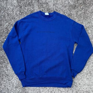 Pode incluir: Uma camisola azul royal com a palavra "provincialism" impressa em preto no peito. A parte de cima de manga comprida é feita de um material macio e tem gola redonda.
